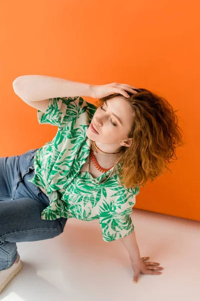 Stilvolle junge rothaarige Frau in Jeans und Bluse mit Blumenmuster, die Kopf und Augen berührt, während sie auf orangefarbenem Hintergrund posiert, trendiges lässiges Sommer-Outfit-Konzept, Jugendkultur — Stockfoto
