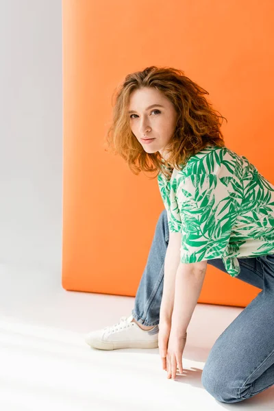 Stylish redhead woman with natural makeup wearing blouse with floral print and jeans while looking at camera on orange background, trendy casual summer outfit concept, Youth Culture — Stock Photo