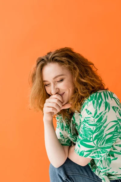 Fröhliche junge rothaarige Frau mit geschlossenen Augen in Bluse mit Blumenmuster und Jeans lacht, während sie isoliert auf orangefarbenem, trendigem Sommeroutfit-Konzept posiert, Jugendkultur — Stockfoto