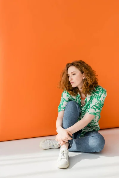 Полная длина молодой рыжеволосой женщины в современной блузке с цветочным узором и джинсами, сидя на сером и оранжевом фоне, модный случайный летний наряд концепции, молодежная культура — стоковое фото