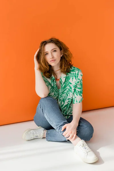 Full length of trendy young redhead woman in blouse with floral pattern, jeans and necklaces looking at camera and sitting on orange background, trendy casual summer outfit concept, Youth Culture — Stock Photo