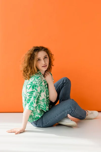 Modelo de pelo rojo joven en blusa moderna con patrón floral y jeans mirando a la cámara mientras está sentado sobre fondo naranja, concepto de atuendo de verano casual de moda, Cultura Juvenil - foto de stock