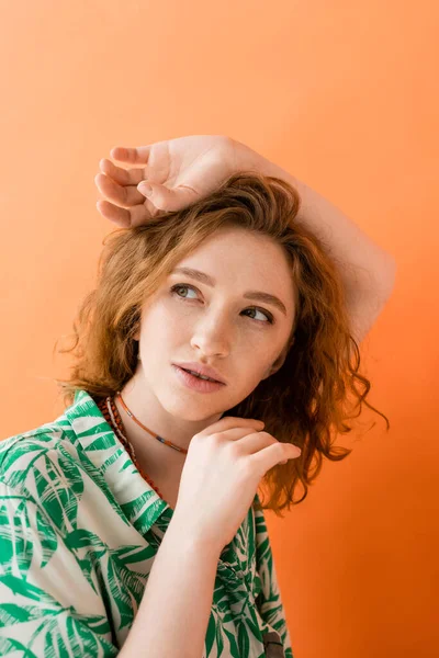 Portrait de femme rousse rêveuse et élégante en chemisier moderne avec motif floral regardant loin et posant isolé sur orange, concept de tenue d'été décontractée à la mode, Culture de la jeunesse — Photo de stock