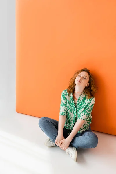 Полная длина расслабленной рыжеволосой женщины в джинсах, современная блузка с цветочным узором и ожерельями, сидя на сером и оранжевом фоне, модная повседневная летняя одежда концепции, молодежная культура — стоковое фото
