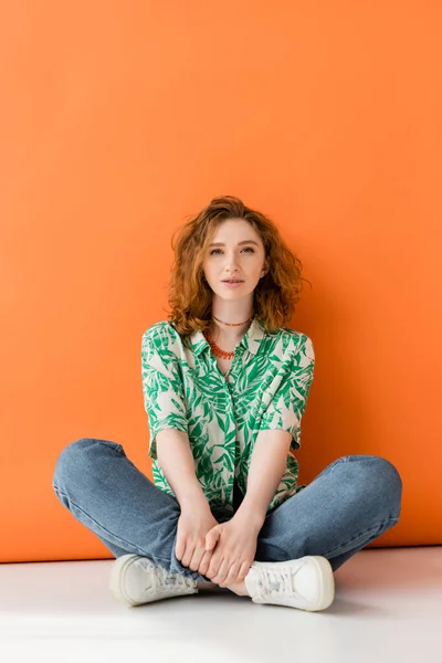 Full length of young red haired woman with natural makeup, jeans and blouse with floral pattern looking at camera on orange background, trendy casual summer outfit concept, Youth Culture — Stock Photo