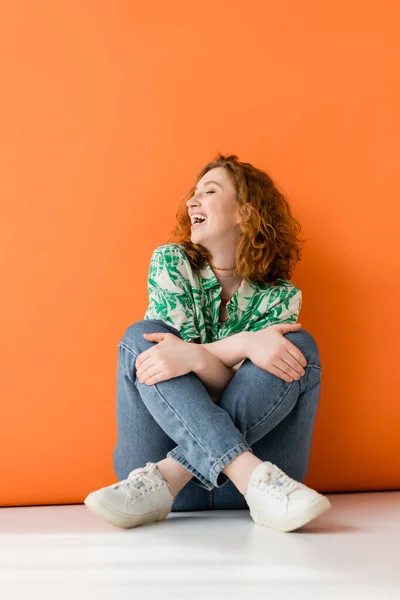 Joyful young red haired model in stylish blouse with floral pattern and modern jeans touching knees and sitting on orange background, trendy casual summer outfit concept, Youth Culture — Stock Photo