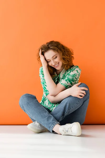 Full length of stylish young red headed woman in blouse with floral pattern and jeans smiling while sitting on orange background, trendy casual summer outfit concept, Youth Culture — Stock Photo