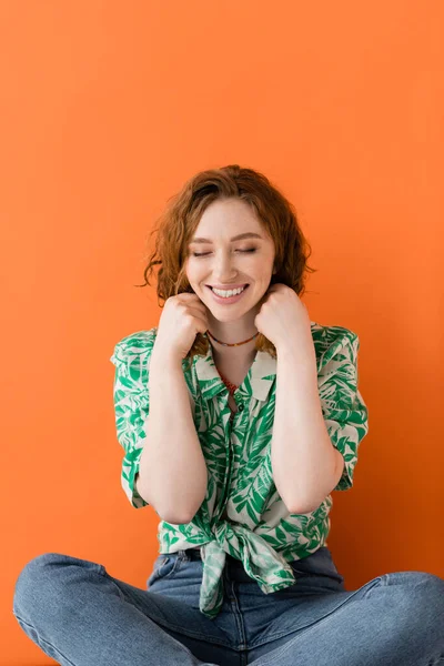 Jovem modelo ruivo alegre em jeans e blusa com padrão floral tocando colares enquanto sentado em fundo laranja, conceito de roupa de verão casual na moda, Cultura Jovem — Fotografia de Stock