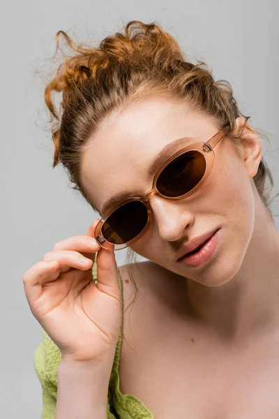 Porträt einer selbstbewussten, rothaarigen und sommersprossigen Frau in grüner Bluse, die Sonnenbrille berührt, während sie posiert und isoliert auf einem grauen, trendigen Sonnenschutzkonzept steht, Jugendkultur — Stockfoto