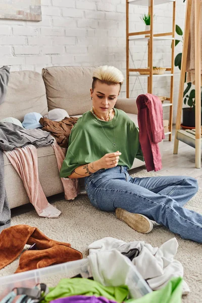 Decluttering process, young and tattooed woman sitting on floor carpet near couch in living room and sorting clothing, trendy hairstyle, tattoo, sustainable living and mindful consumerism concept — Stock Photo