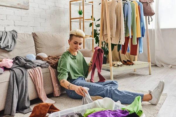 Tattooed woman sorting wardrobe items while sitting on floor in living room near couch and rack with clothes, decluttering and reducing, sustainable living and mindful consumerism concept — Stock Photo
