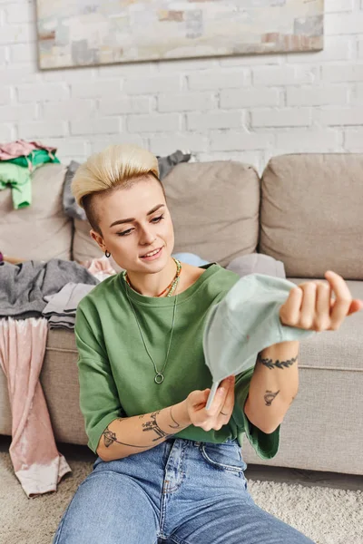 Wardrobe items decluttering, young and cheerful woman with tattoo and trendy hairstyle holding cap near couch with clothes in living room, sustainable living and mindful consumerism concept — Stock Photo