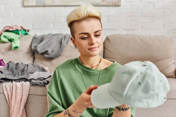 Smiling woman looking at cap while sorting wardrobe items near couch with clothes at home, positive emotion, trendy hairstyle, tattoo, sustainable living and mindful consumerism concept — Stock Photo