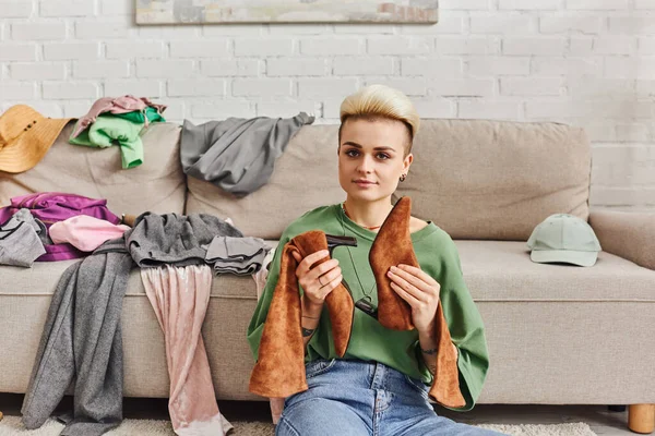 Sorting garments, young woman with trendy hairstyle and tattoo holding suede boots, looking at camera near garments on couch in modern living room, sustainable living and mindful consumerism concept — Stock Photo