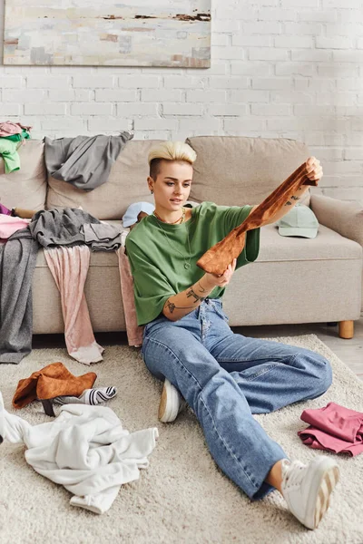 Tattooed woman in casual style clothes holding suede boot while sorting belongings and thrift store finds near couch on floor in living room, sustainable living and mindful consumerism concept — Stock Photo