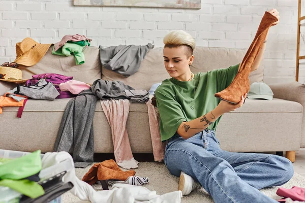 Young woman sitting on floor with suede boot near clothes and couch at home, sorting and reducing wardrobe, trendy hairstyle, tattoo, sustainable living and mindful consumerism concept — Stock Photo