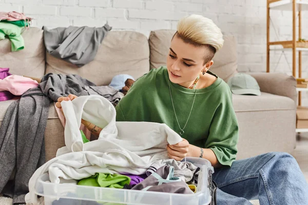 Young woman with tattoo and trendy hairstyle sorting second-hand clothes in plastic container near couch in modern living room, sustainable living and mindful consumerism concept — Stock Photo