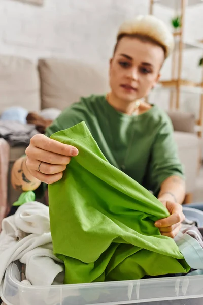 Young tattooed woman reducing wardrobe, sorting clothes and holding green garment in living room at home, blurred background, sustainable living and mindful consumerism concept — Stock Photo