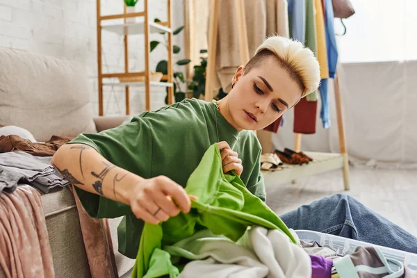 Молодая, стильная и татуированная женщина с зеленой одеждой в современной гостиной у себя дома, сортировка и разглаживание, устойчивая жизнь и внимательный концепция потребительства — стоковое фото