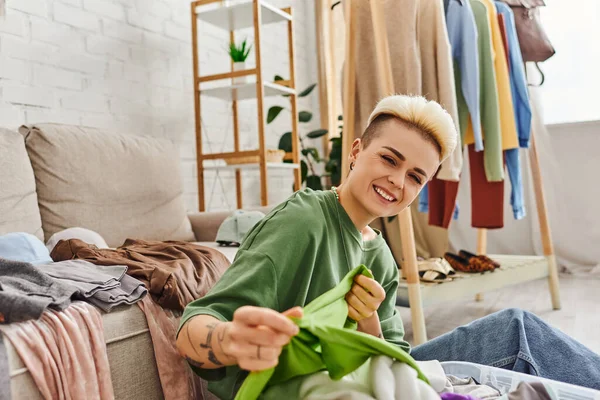 Young excited woman sorting second-hand clothes and holding green garment near couch and rack at home, trendy hairstyle, tattoo, looking at camera, sustainable living and mindful consumerism concept — Stock Photo