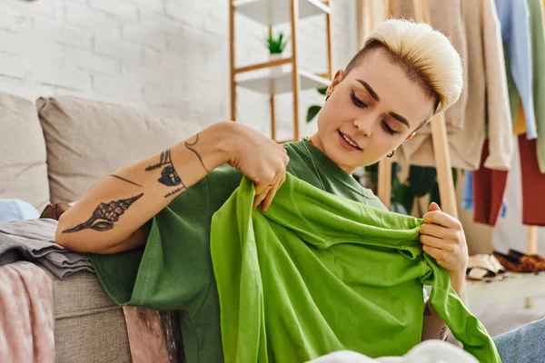 Довольная женщина, держащая зеленую одежду рядом с диваном в гостиной, в то время как раздевание одежды дома, модная прическа, татуировка, устойчивая жизнь и внимательный концепция потребительства — стоковое фото