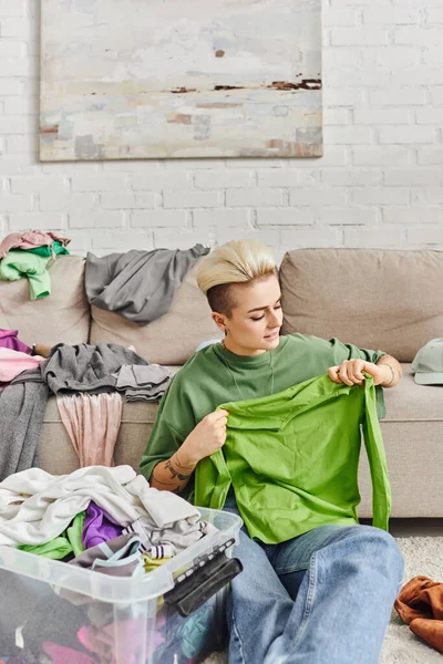 Young woman with trendy hairstyle and tattoo, holding green garment near plastic container and couch with clothes, sorting, conscious decluttering, sustainable living and mindful consumerism concept — Stock Photo