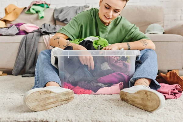 Smiling and tattooed woman sitting on floor near plastic container with thrift store finds, decluttering process, clothes sorting, sustainable living and mindful consumerism concept — Stock Photo