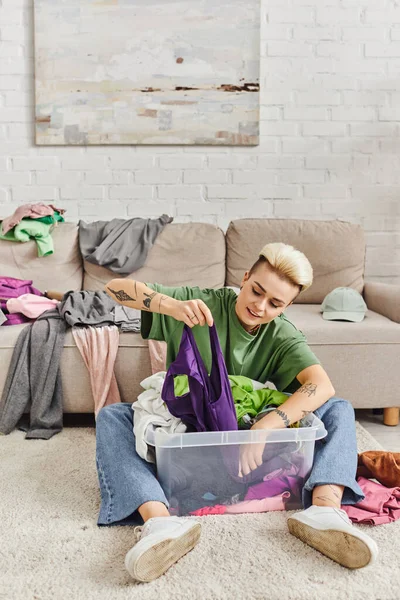 Stylish tattooed woman with trendy hairstyle sorting clothes in plastic container on floor near couch with wardrobe items in modern living room, sustainable living and mindful consumerism concept — Stock Photo
