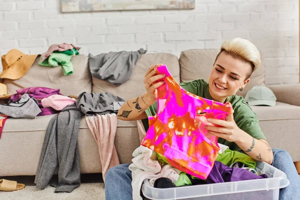 Young woman with cheerful smile sorting thrift store finds, holding colorful top near plastic container and couch, trendy hairstyle, tattoo, sustainable living and mindful consumerism concept — Stock Photo