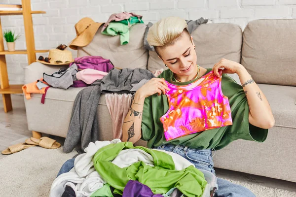 Mujer sonriente sosteniendo la parte superior colorida mientras decluttering artículos de armario cerca del sofá con ropa en la sala de estar moderna, peinado de moda, tatuaje, vida sostenible y concepto de consumismo consciente - foto de stock