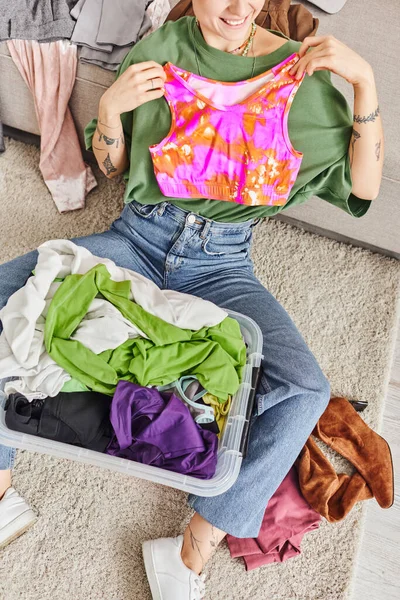 Cropped view of woman with happy smile and tattoo holding colorful top near plastic container with clothing, decluttering process, sustainable living and mindful consumerism concept, top view — Stock Photo