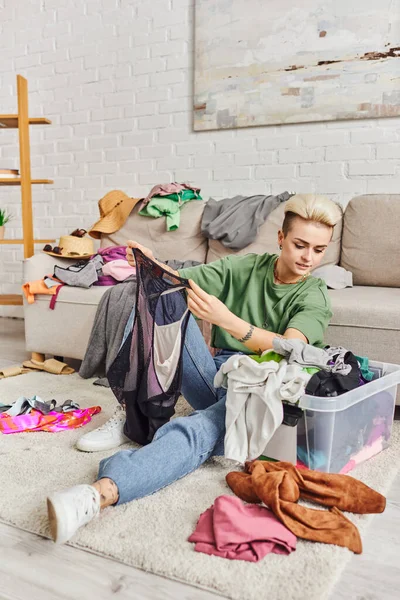 Casually styled woman holding bodysuit on floor near plastic container with clothes, decluttering wardrobe items, trendy hairstyle, tattoo, sustainable living and mindful consumerism concept — Stock Photo