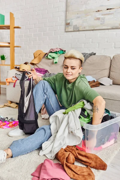 Tattooed woman sitting on floor near plastic container and sorting second-hand clothes near couch at home, trendy hairstyle, pleased smile, sustainable living and mindful consumerism concept — Stock Photo