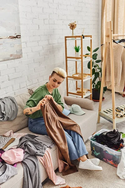 Jovem mulher tatuada segurando calças de couro enquanto sentado no sofá perto de roupas, prateleiras e plantas na sala de estar moderna, processo de decluttering, vida sustentável e conceito de consumismo consciente — Fotografia de Stock