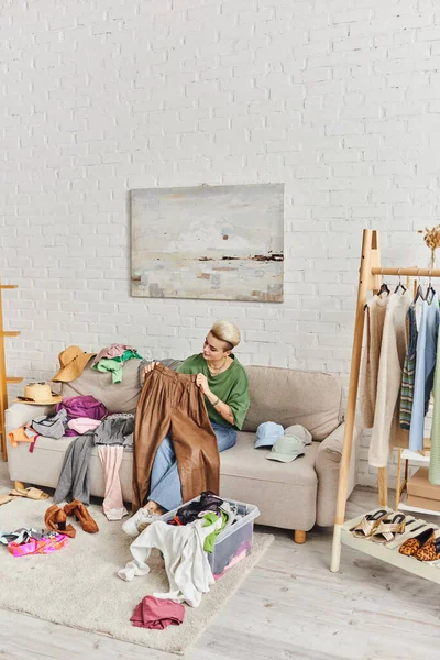 Tätowierte Frau sitzt auf Couch neben Kleiderschrank, hält Lederhose, sortiert Kleidung, modernes Wohnzimmer, Kleiderständer, nachhaltiges Leben und achtsames Konsumkonzept — Stockfoto