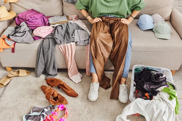 Vue partielle d'une femme tatouée et élégante assise sur un canapé avec un pantalon en cuir pendant le déballage des vêtements à la maison dans le salon, un mode de vie durable et un concept de consumérisme conscient — Photo de stock