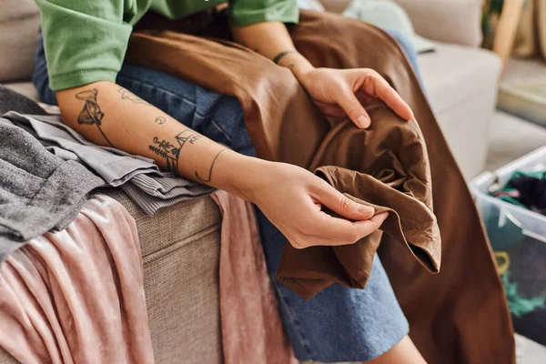 Partial view of young and stylish woman with tattooed arms holding leather pants, sitting on couch at home and sorting clothes to reduce wardrobe, sustainable living and mindful consumerism concept — Stock Photo