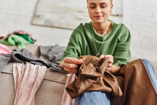 Low angle view of young woman holding stylish leather pants while sitting on couch in living room and sorting clothes at home,  blurred background, sustainable living and mindful consumerism concept — Stock Photo