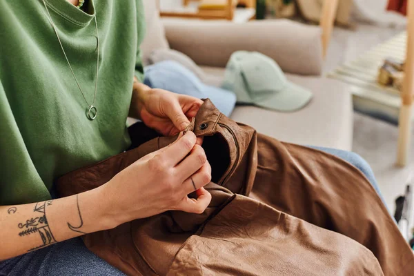 Vista recortada de la joven mujer tatuada abotonar pantalones de cuero mientras está sentado en el sofá cerca de artículos de armario durante el proceso de decluttering, vida sostenible y concepto de consumismo consciente - foto de stock