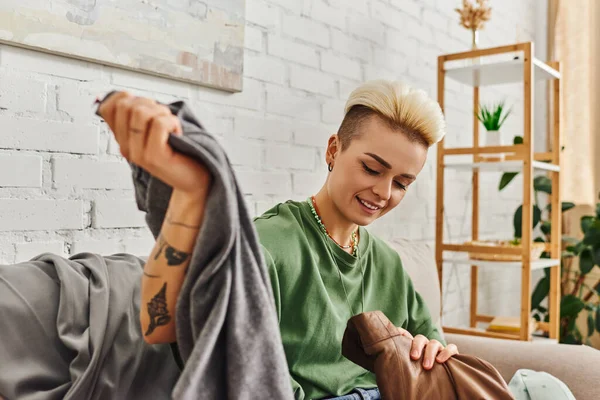 Carefree woman with tattoo and trendy hairstyle sorting pre-loved items and second-hand clothing in modern living room, wardrobe reducing, sustainable living and mindful consumerism concept — Stock Photo