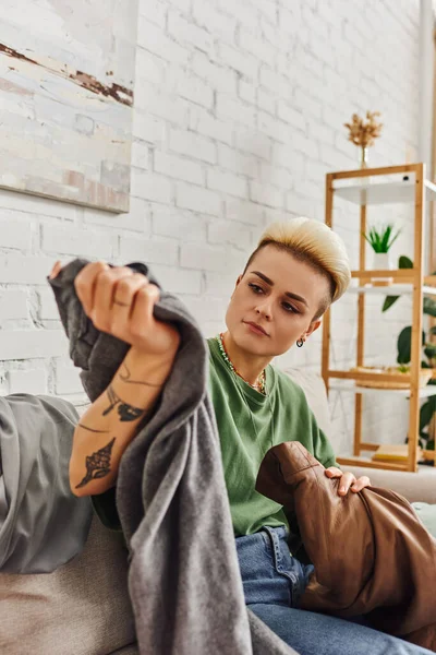 Tattooed woman with trendy hairstyle sorting clothes and reducing wardrobe items on couch in living room near rack with green plants at home, sustainable living and mindful consumerism concept — Stock Photo