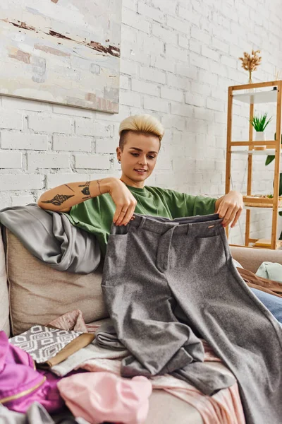 Giovane e sorridente donna tatuata che tiene i pantaloni grigi mentre siede sul divano vicino agli indumenti in salotto moderno, ricerca di negozi di risparmio, concetto di vita sostenibile e consumismo consapevole — Foto stock