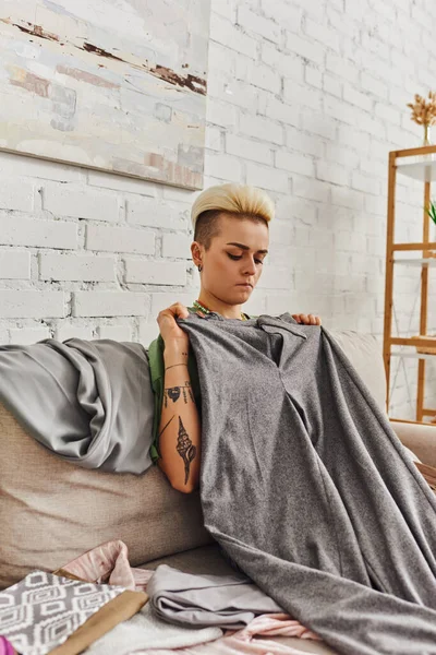 Thoughtful young woman with trendy hairstyle and tattoo sitting on couch near wardrobe items and holding grey pants, clothes sorting, decluttering, sustainable living and mindful consumerism concept — Stock Photo