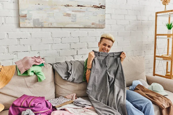 Overjoyed tattooed woman holding grey pants and looking at camera near clothes on couch in living room, sorting wardrobe items, home decluttering, sustainable living and mindful consumerism concept — Stock Photo