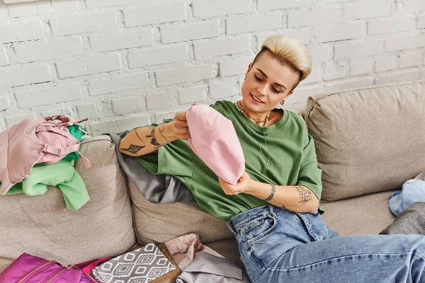 Joyful and tattooed woman with trendy hairstyle looking at cap while sitting on couch, sorting clothes and decluttering wardrobe, sustainable living and mindful consumerism concept — Stock Photo
