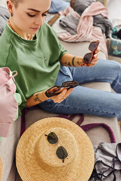 Високий кут зору стильна і татуйована жінка тримає сонцезахисні окуляри біля солом'яного капелюха і предмети гардеробу на дивані вдома, сортує одяг, стійке життя і продуману концепцію споживання — стокове фото