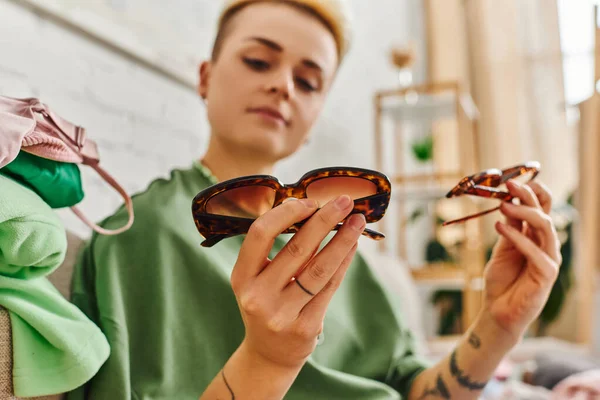 Low angle view of young tattooed woman holding trendy sunglasses while sorting and decluttering clothing and pre-loved items, blurred background, sustainable living and mindful consumerism concept — Stock Photo