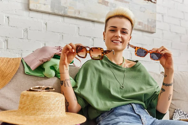 Happy young woman sitting on couch near straw hat and clothing while showing stylish sunglasses, trendy hairstyle, tattoo, sustainable living and mindful consumerism concept — Stock Photo