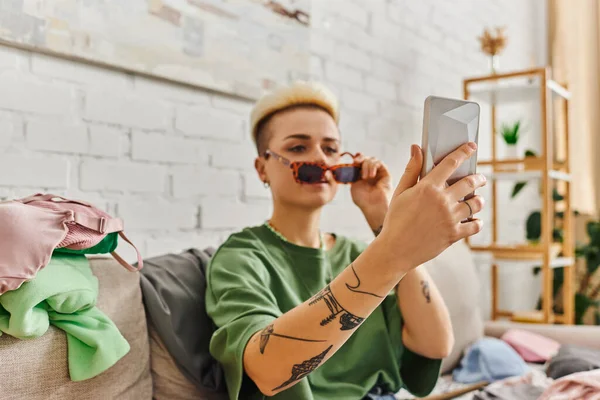 Femme tatouée avec coiffure à la mode prenant selfie dans des lunettes de soleil élégantes pour l'échange sur le marché en ligne, effets decluttering, vie durable et concept de consumérisme conscient — Photo de stock