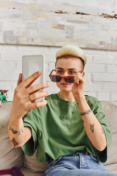 Online internet swap, conscious decluttering, young tattooed woman with trendy hairstyle taking selfie in fashionable sunglasses on smartphone, sustainable living and mindful consumerism concept — Stock Photo